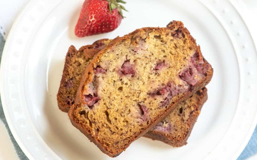 Two slices of strawberry banana bread are stacked on a white plate, with a whole strawberry placed beside them.