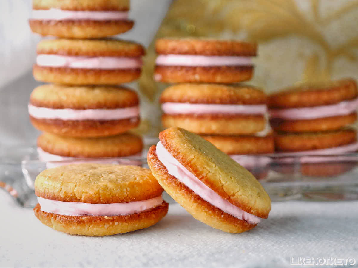 A stack of sandwich cookies with pink cream filling is displayed, with two cookies in the foreground and more cookies stacked in the background.