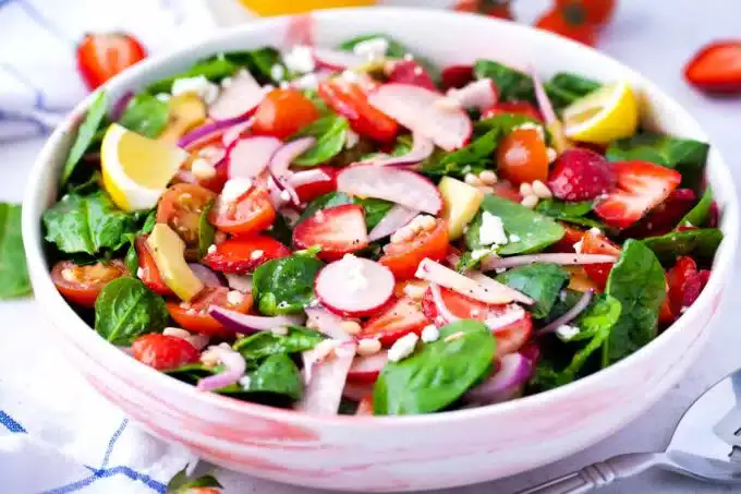 A bowl of fresh salad with spinach, cherry tomatoes, strawberries, radishes, red onions, lemon wedges, feta cheese, and pine nuts.