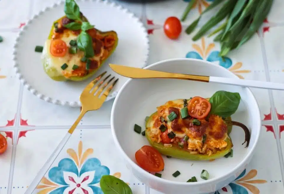 A stuffed bell pepper filled with cheese, vegetables, and topped with tomato slices and basil, served on a white plate and a bowl with a gold fork and knife.