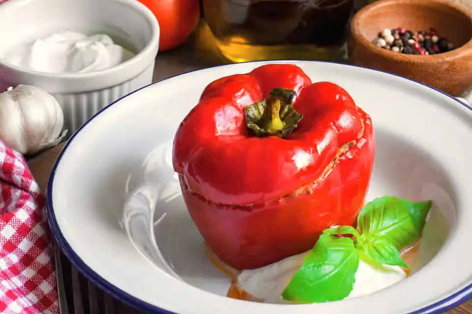 A stuffed red bell pepper sits on a plate with a dollop of white sauce and fresh basil garnish, surrounded by various ingredients in the background.