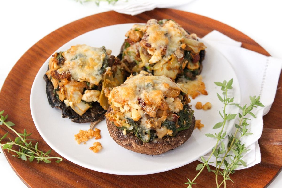 Three stuffed portobello mushrooms topped with melted cheese are served on a white plate, garnished with sprigs of fresh herbs.