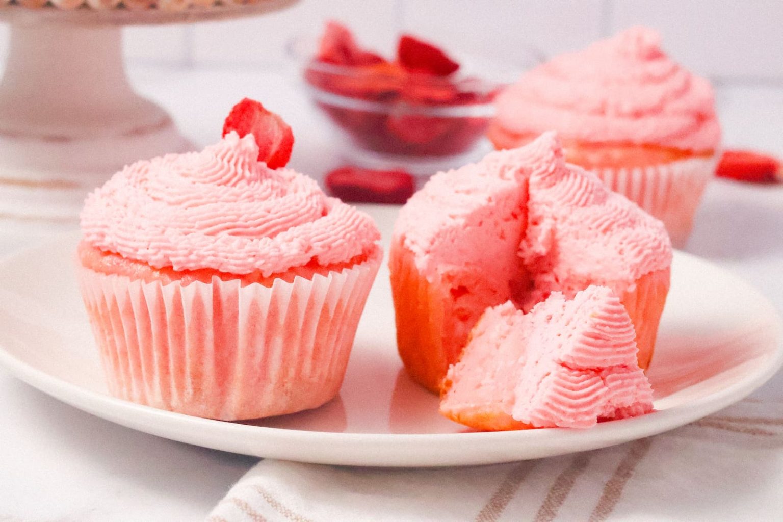 A plate with three pink frosted cupcakes, one of which is partially cut to show the inside, sits on a white surface with a bowl of strawberries in the background.