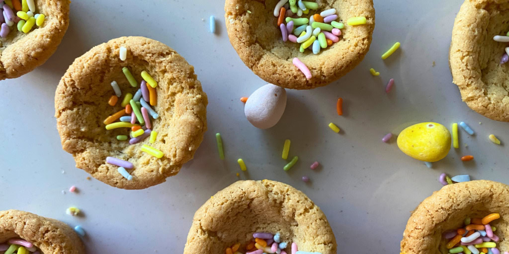 Cookies with hollow centers filled with colorful sprinkles are arranged on a white surface, with a few small candy eggs scattered nearby.