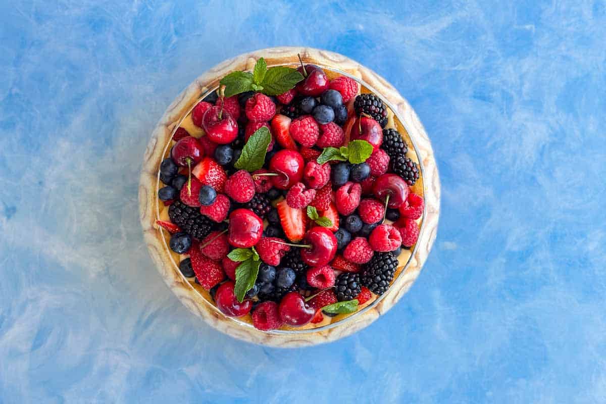 A round fruit tart topped with cherries, raspberries, blueberries, strawberries, blackberries, and mint leaves on a blue textured surface.