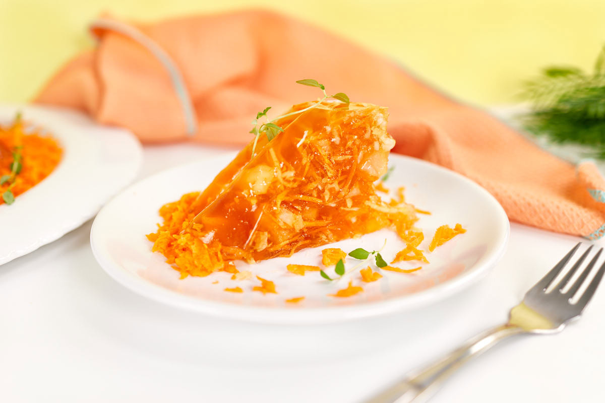 A slice of carrot cake topped with orange glaze and herb garnish is served on a white plate, with a fork and an orange napkin in the background.