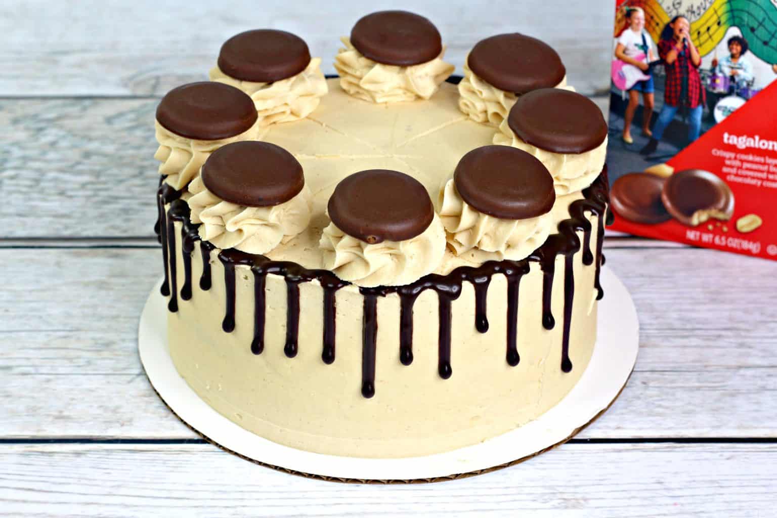 A round frosted cake with chocolate drip, decorated with swirls of frosting and chocolate-covered cookies on top. A box of Tagalongs cookies is partially visible in the background.