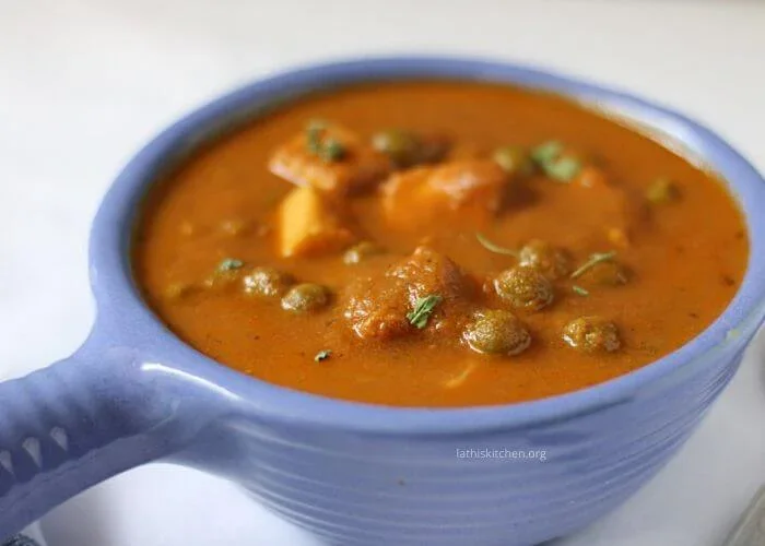 A blue bowl filled with a thick, orange-brown curry containing pieces of paneer and green peas, garnished with herbs.