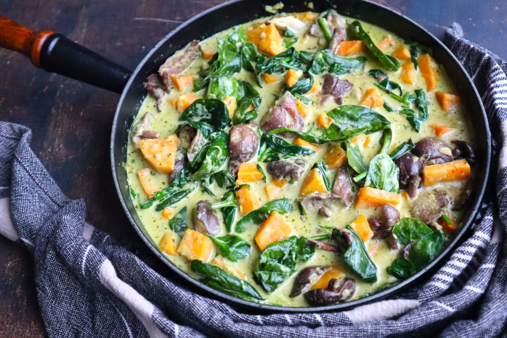 A skillet filled with curry containing chunks of sweet potato, pieces of meat, fresh spinach leaves, and a creamy yellow sauce.