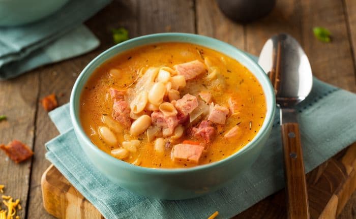 A bowl of creamy ham and bean soup with chunks of ham and white beans, placed on a blue napkin with a spoon beside it on a wooden table.