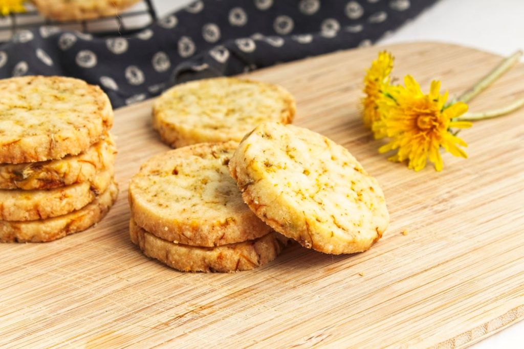 Round shortbread cookies with visible herb flecks are arranged on a wooden board next to two yellow dandelion flowers.