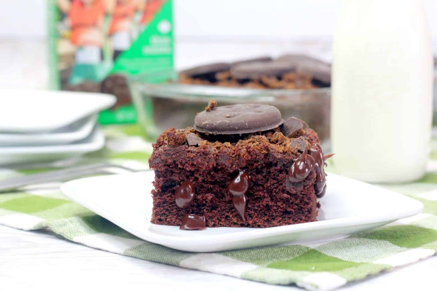 A slice of chocolate cake topped with chocolate pieces and a chocolate-covered cookie sits on a white plate, with a bottle of milk in the background.