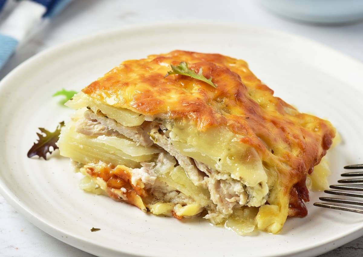 A close-up of a slice of baked potato casserole with melted cheese and chicken layers on a white plate, garnished with a small piece of herb.