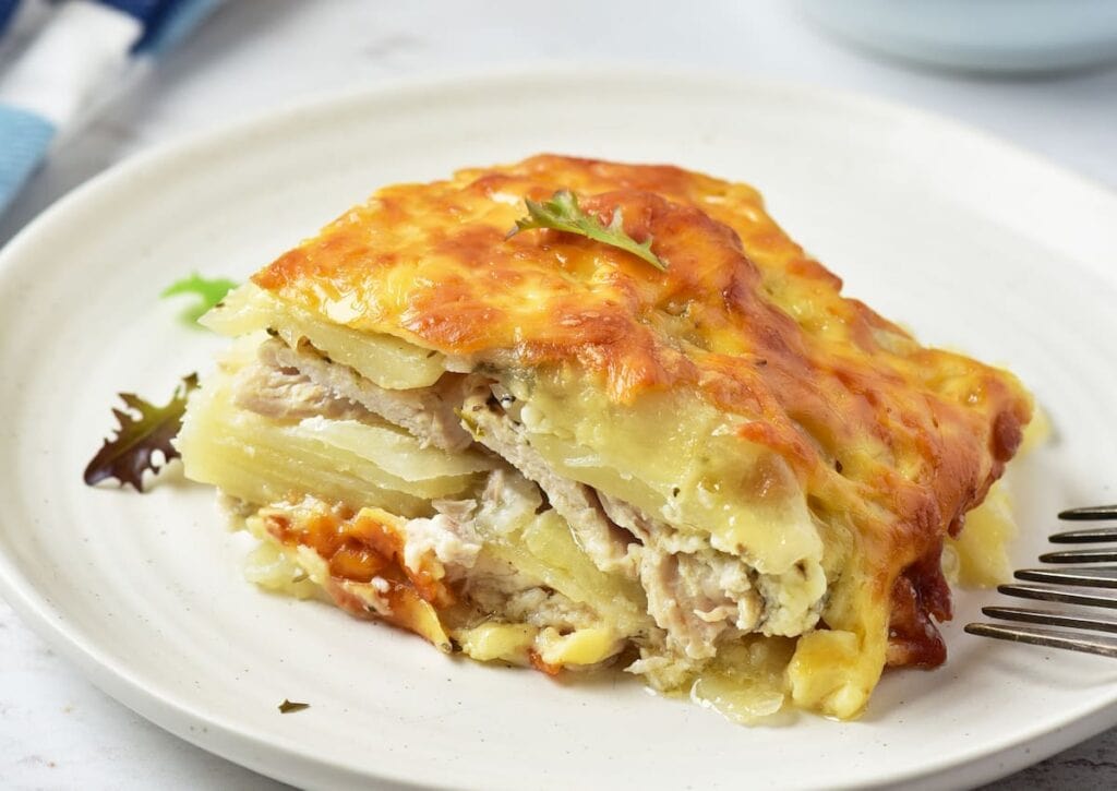 A slice of creamy potato and chicken casserole topped with melted cheese, served on a white plate with a small garnish and a fork on the side.