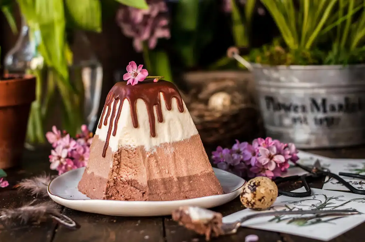 A pyramid-shaped layered chocolate and vanilla dessert with chocolate glaze, garnished with pink flowers, on a white plate surrounded by spring decor and quail eggs.