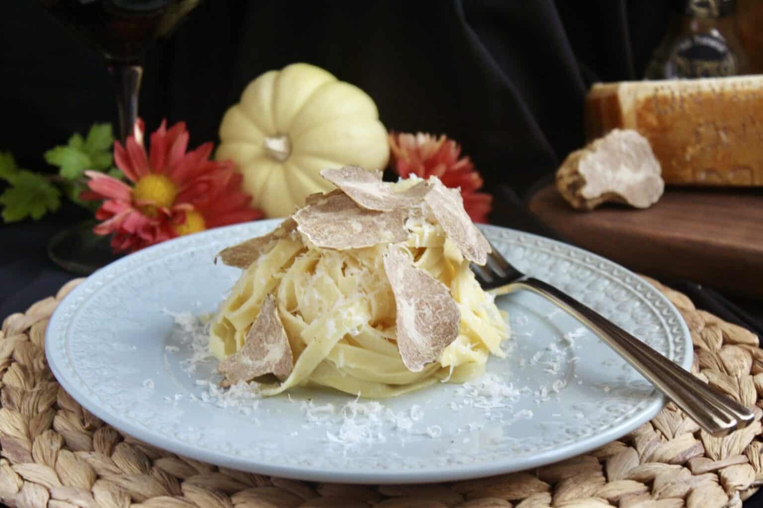 A plate of tagliatelle pasta topped with shaved truffles and grated cheese, set on a woven placemat with flowers, a small pumpkin, and cheese in the background.