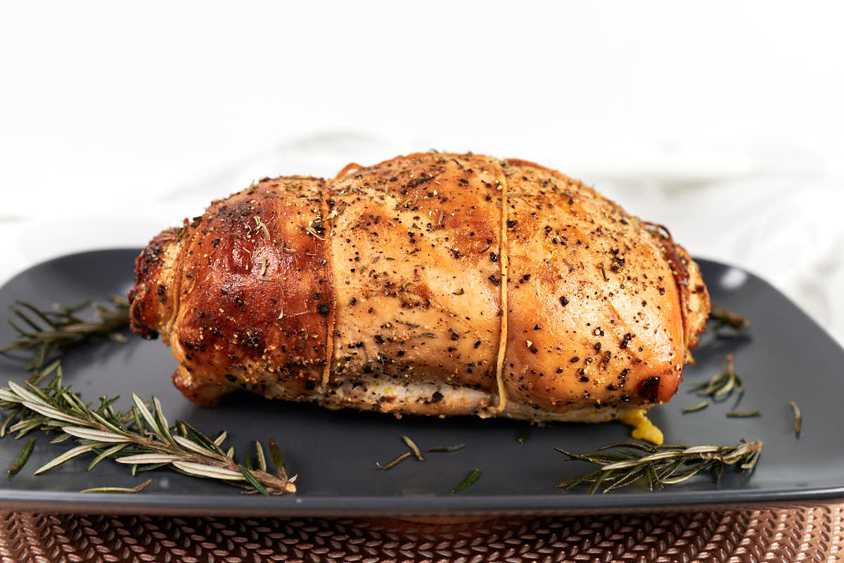 A cooked, seasoned rolled roast tied with string sits on a dark plate, garnished with sprigs of rosemary.