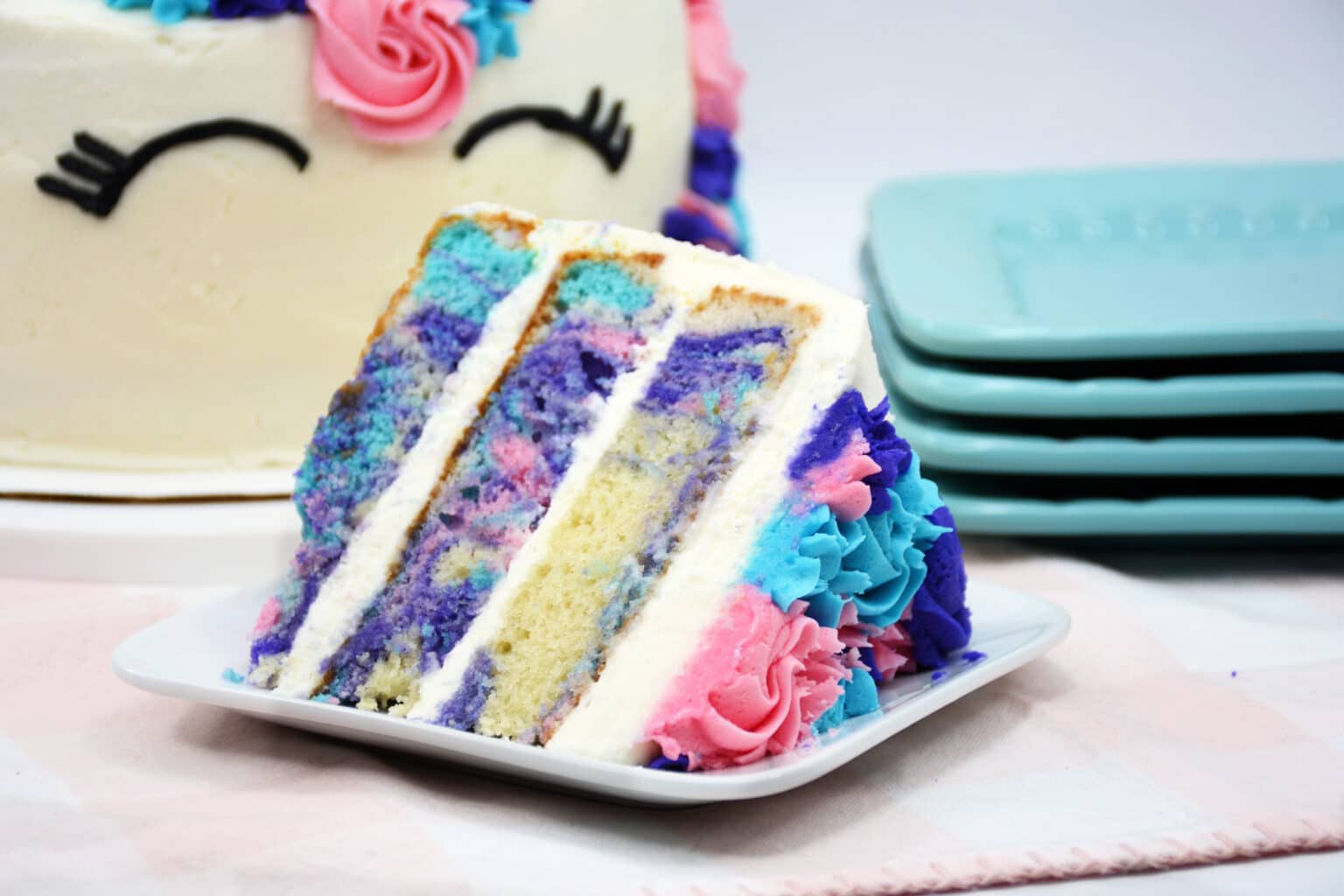A slice of colorful, marbled layer cake with white icing and blue, pink, and purple decorations on a white plate, with part of the whole cake and stacked plates in the background.