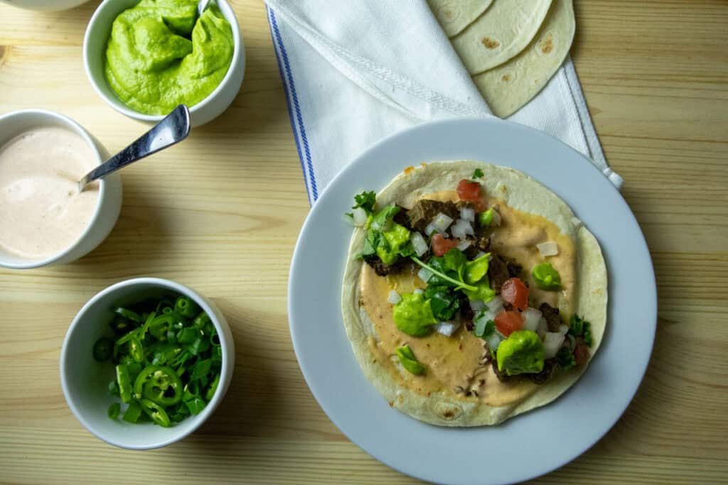 A plate with a tortilla topped with beans, vegetables, and sauces sits beside bowls of guacamole, sliced green onions, and creamy sauce on a wooden table.