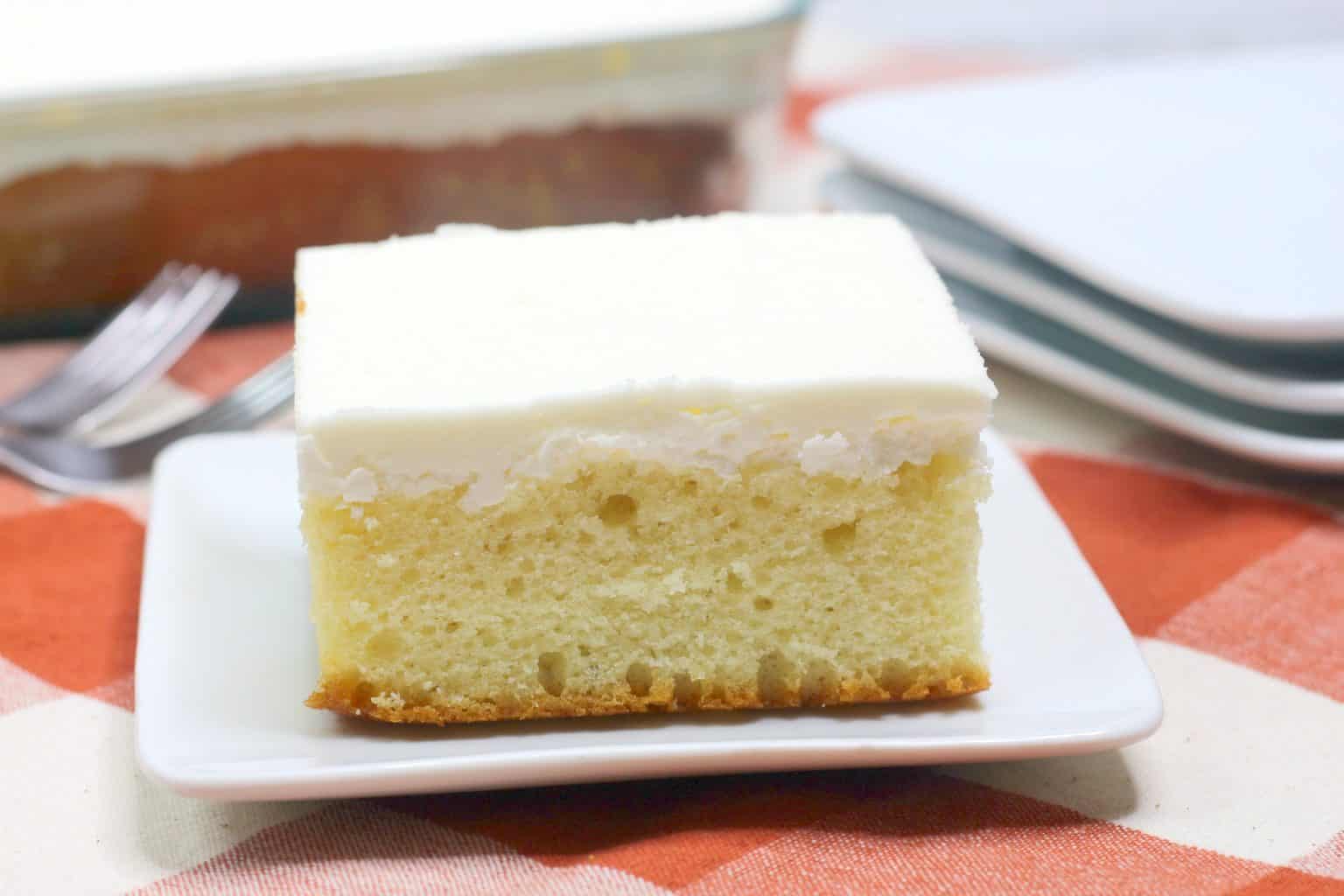 A slice of yellow cake with white frosting on a white plate, with a pan of cake, forks, and stacked plates in the background.