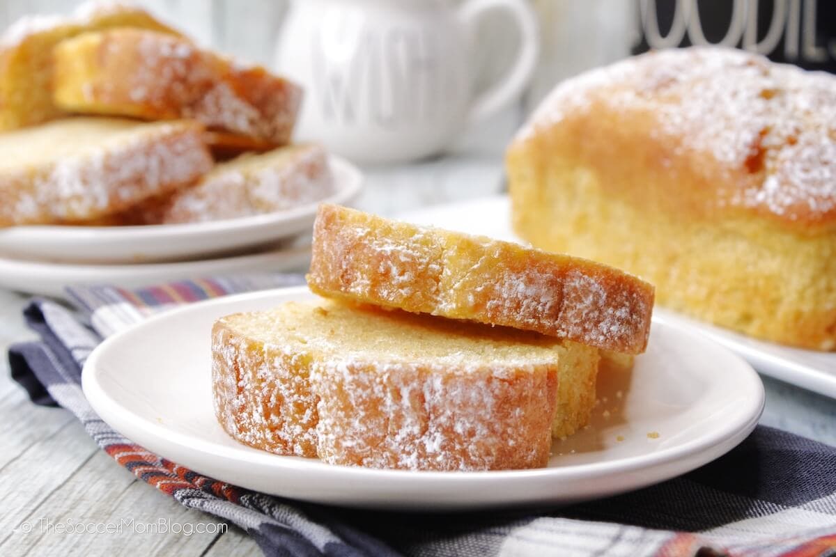 Two slices of powdered sugar-topped pound cake rest on a white plate, with more slices and a loaf in the background.