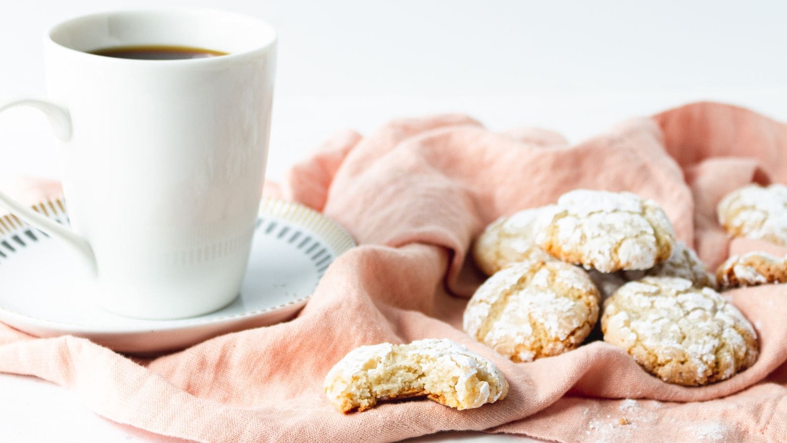 A white cup of coffee on a saucer sits next to several powdered sugar cookies on a pink cloth; one cookie has a bite taken out of it.