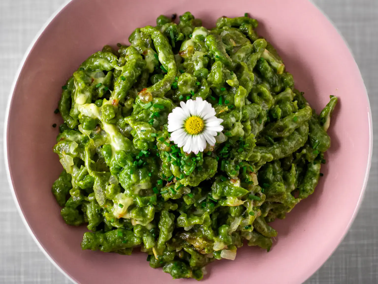 A pink bowl filled with green pasta garnished with chopped herbs and a white daisy flower on top.