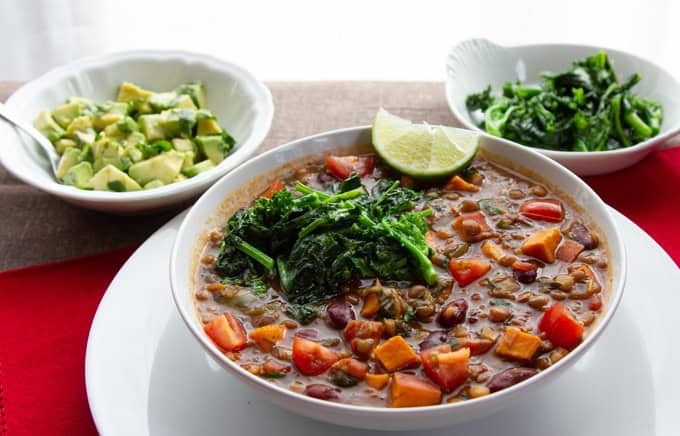 A bowl of vegetable stew with greens and lime wedges, served with sides of chopped avocado and sautéed greens on a white plate.