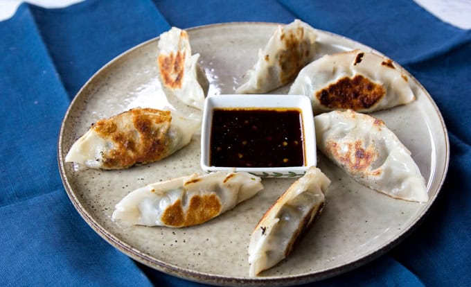 A plate with seven pan-fried dumplings arranged in a circle around a small square dish of soy dipping sauce, set on a blue cloth.