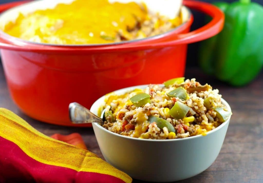 A bowl of cheesy casserole with rice, ground meat, and green bell peppers sits in front of a large red baking dish filled with the same casserole.