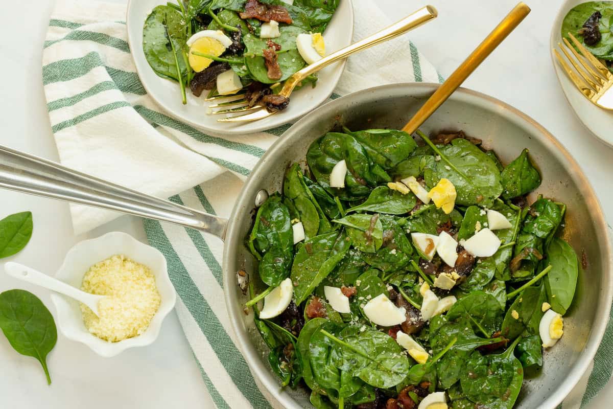 A skillet with spinach salad topped with chopped hard-boiled eggs and bacon, next to a plate of salad and a small bowl of grated cheese.