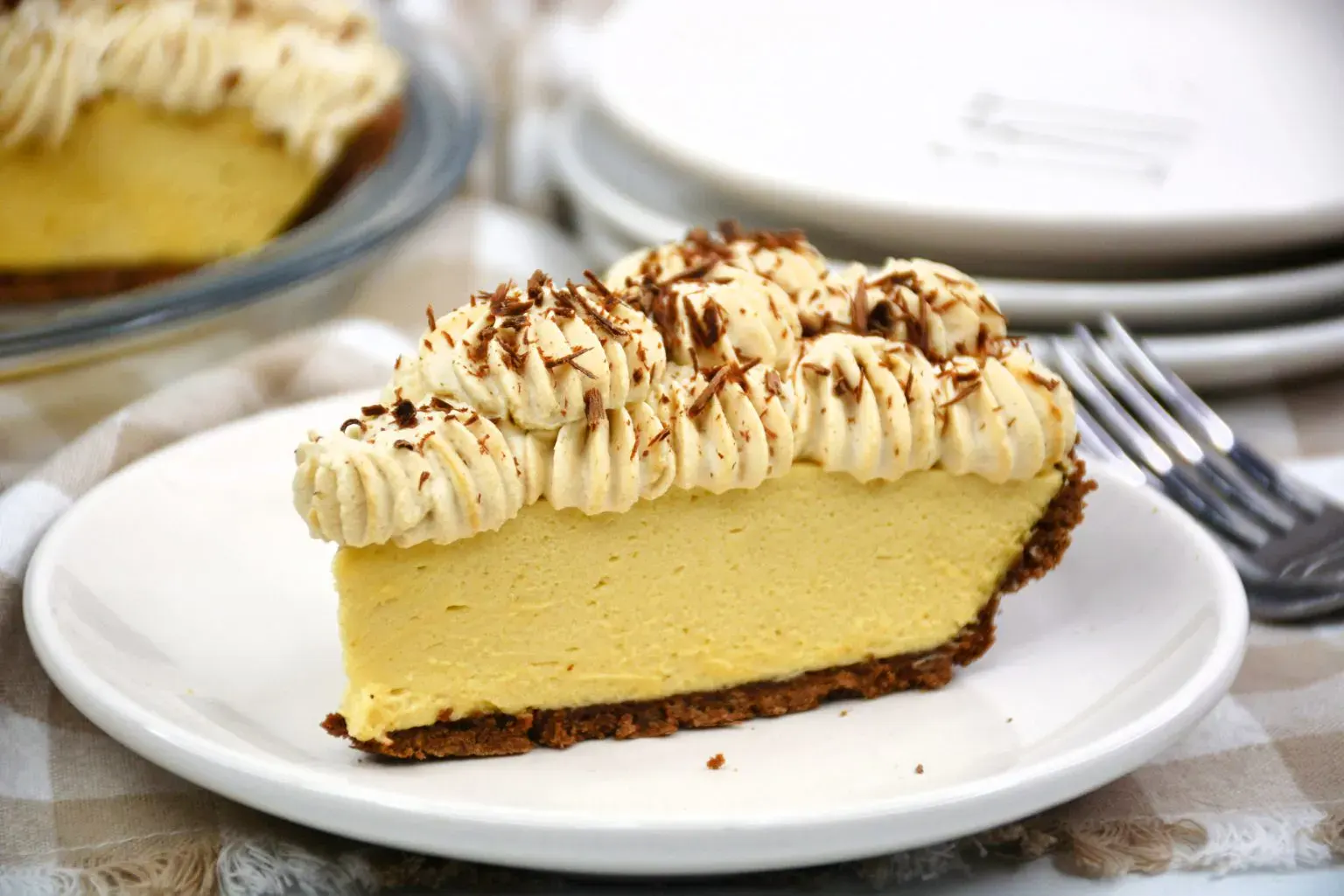 A slice of custard pie with whipped cream topping and chocolate shavings on a white plate, with stacked plates and forks in the background.
