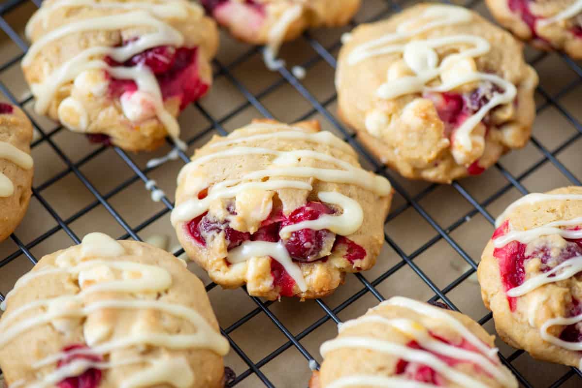 Cookies with visible chunks of cranberries and white chocolate, drizzled with white icing, cooling on a black wire rack.