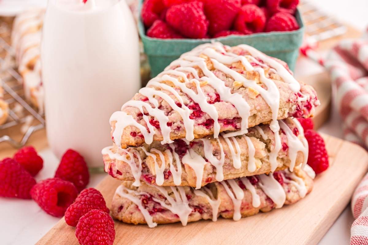 Three raspberry scones with white icing stacked on a wooden board, fresh raspberries and a bottle of milk in the background.