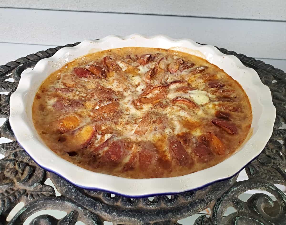 A baked dessert featuring sliced peaches arranged in a circular pattern in a white pie dish, resting on a decorative metal table.