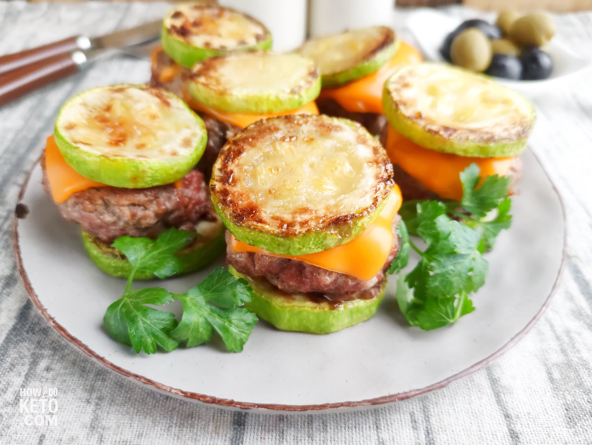 A plate of mini burgers with grilled zucchini slices as buns, cheddar cheese, ground beef patties, and parsley garnish.