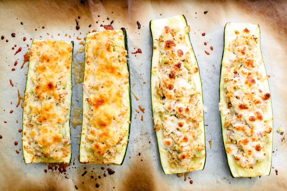 Four baked zucchini halves stuffed with a cheese and breadcrumb mixture are arranged on a parchment-lined baking sheet.
