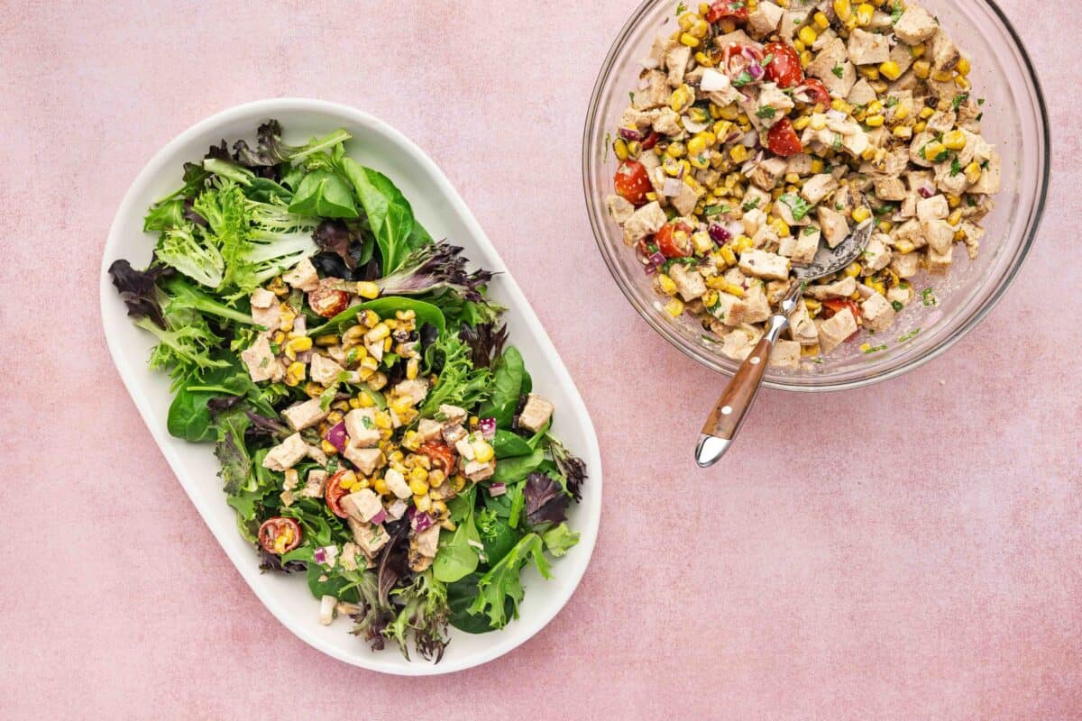 A bowl of mixed salad greens topped with a corn, tomato, and chicken salad. A larger bowl of the same chicken salad with a serving spoon sits beside it on a pink surface.