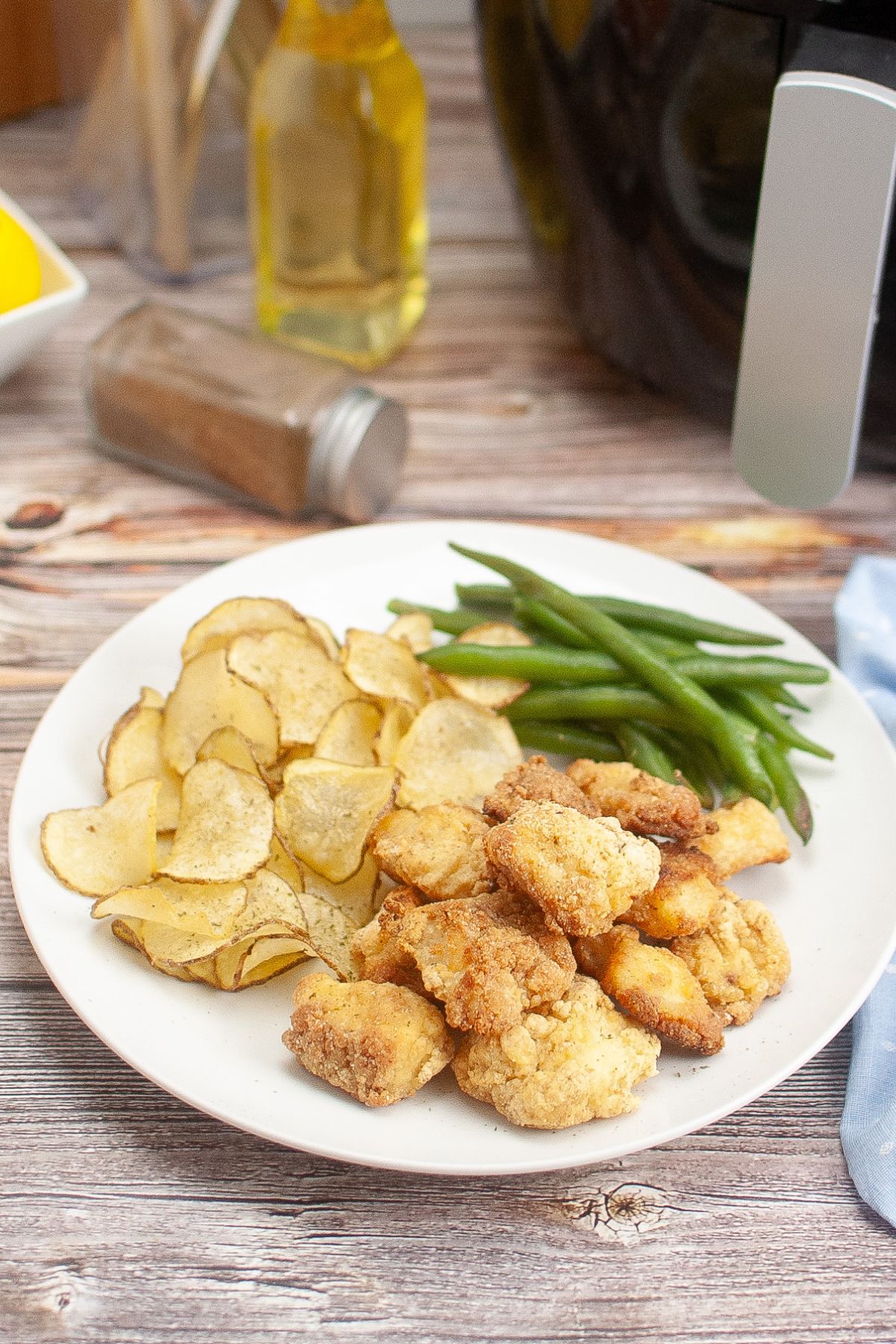 A plate with sliced potato chips, breaded chicken pieces, and green beans on a wooden table, with a spice jar, oil bottle, and air fryer in the background.