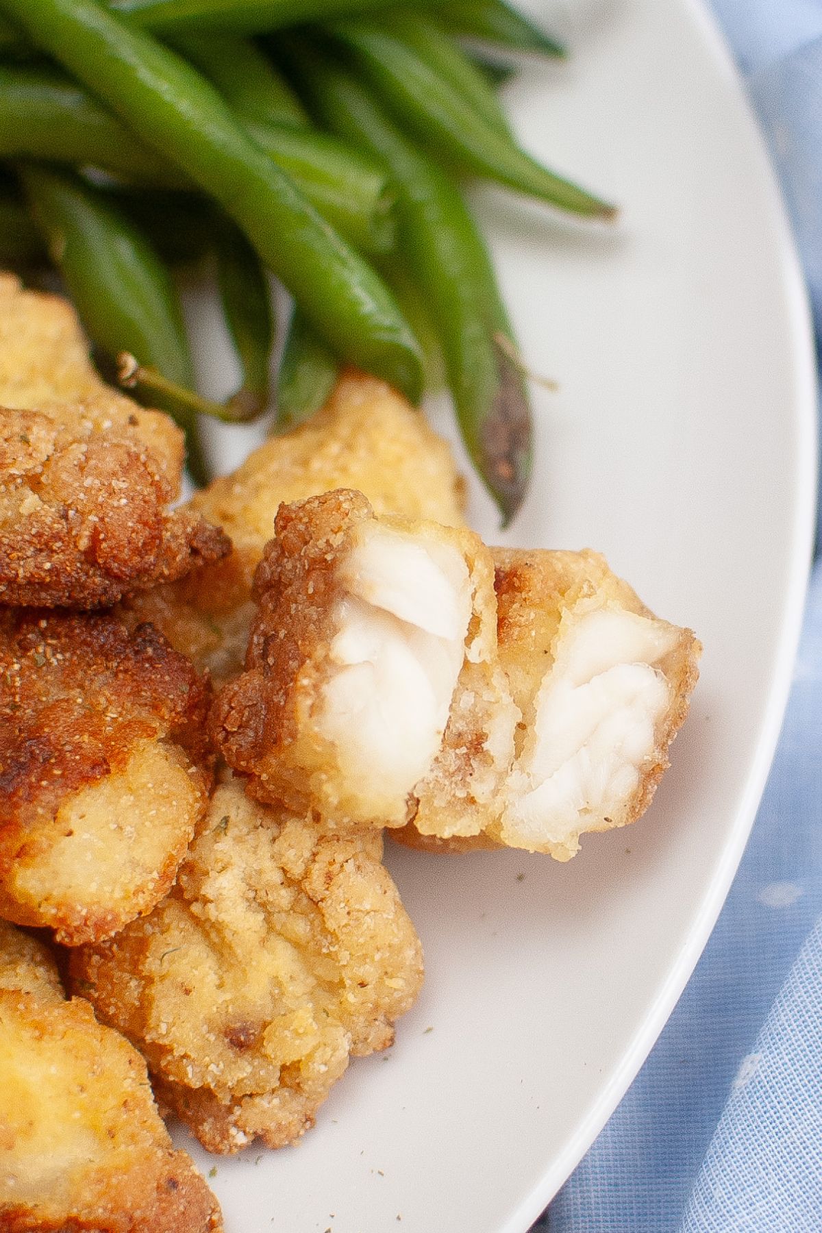 A plate with golden-brown fried fish pieces and green beans, with one piece of fish cut open to show the white, flaky interior.