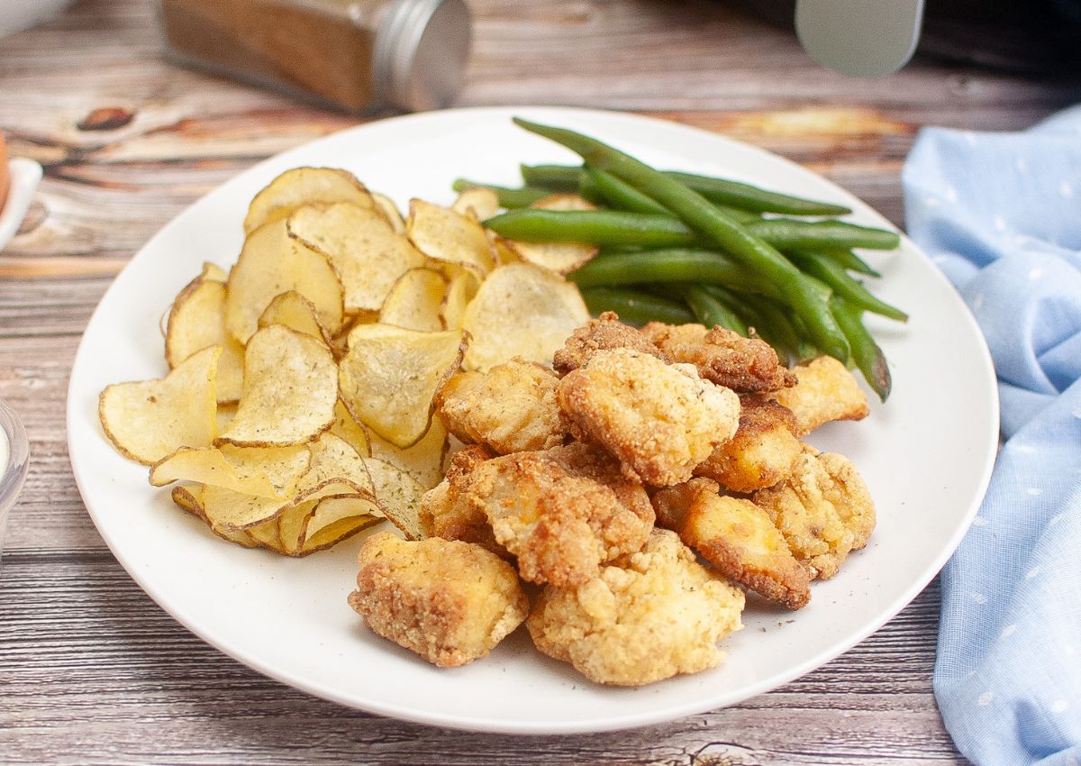 A plate with fried chicken pieces, thinly sliced potato chips, and steamed green beans on a wooden table.