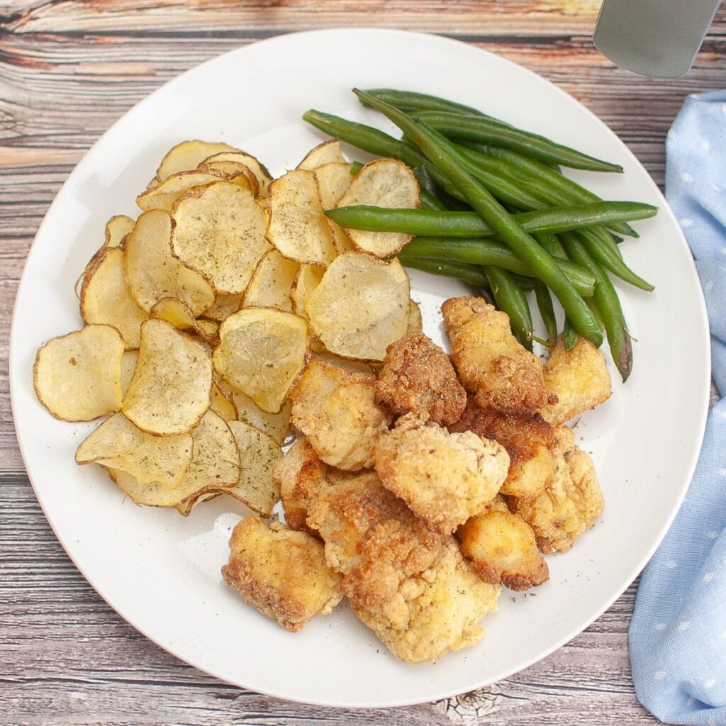 A white plate with breaded chicken pieces, thin potato slices, and steamed green beans, placed on a wooden surface.