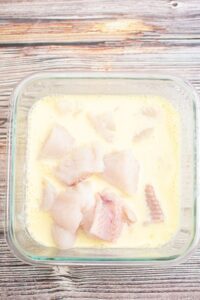 Raw fish pieces soaking in a light yellow liquid, likely an egg or milk mixture, inside a square glass container on a wooden surface.