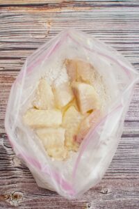 Raw fish pieces coated with flour in a clear plastic bag, placed on a wooden surface.