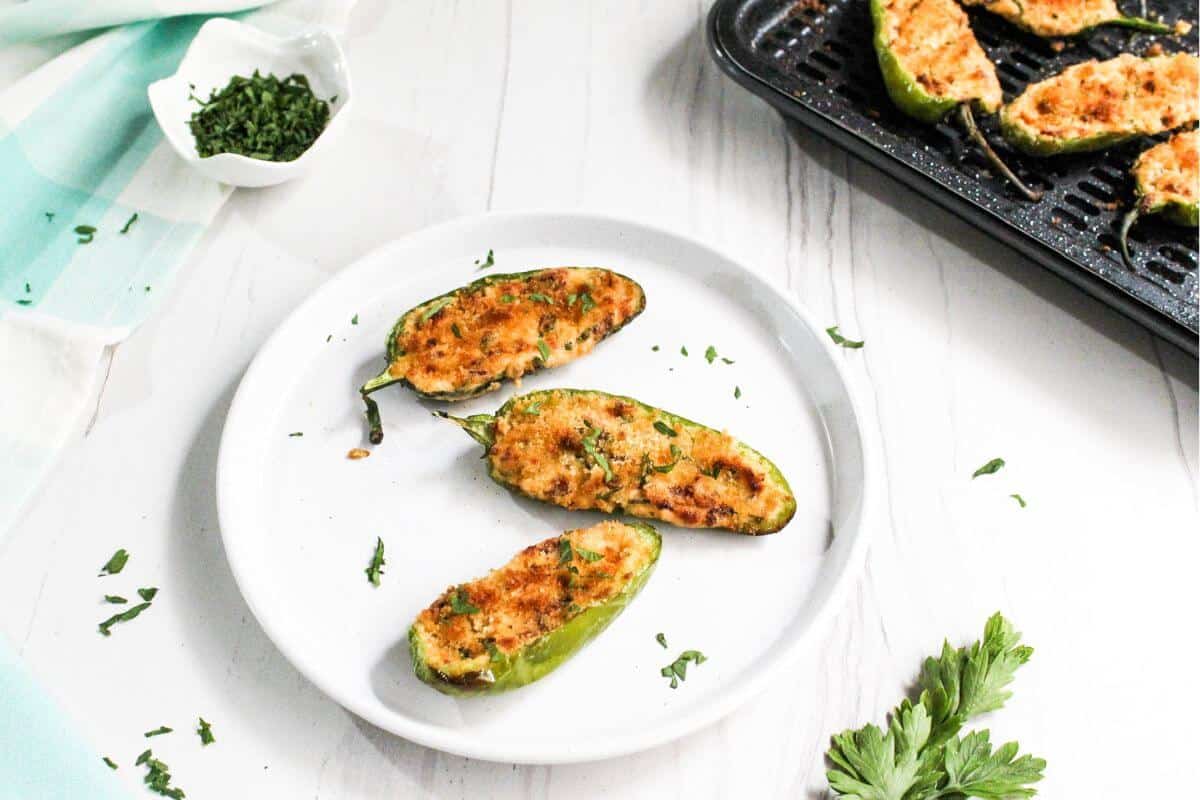 Three baked stuffed jalapeño halves on a white plate, garnished with chopped herbs, with more jalapeños on a baking tray and herbs in a small bowl nearby.