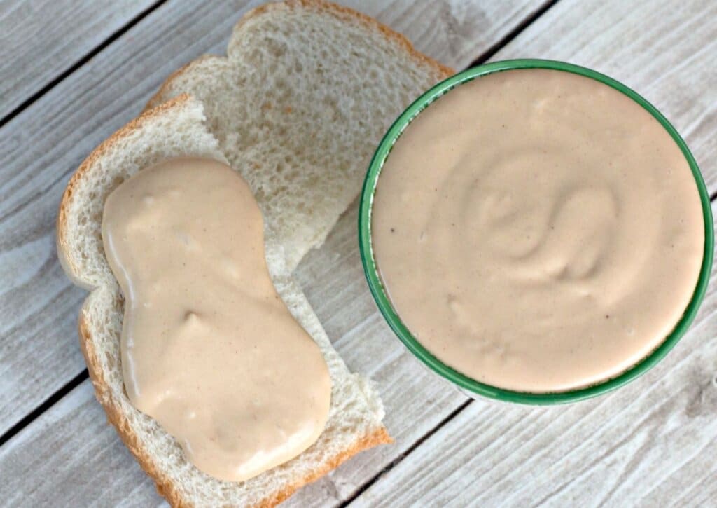 A slice of white bread partially covered with creamy peanut butter sits next to a green bowl filled with more peanut butter on a wooden surface.