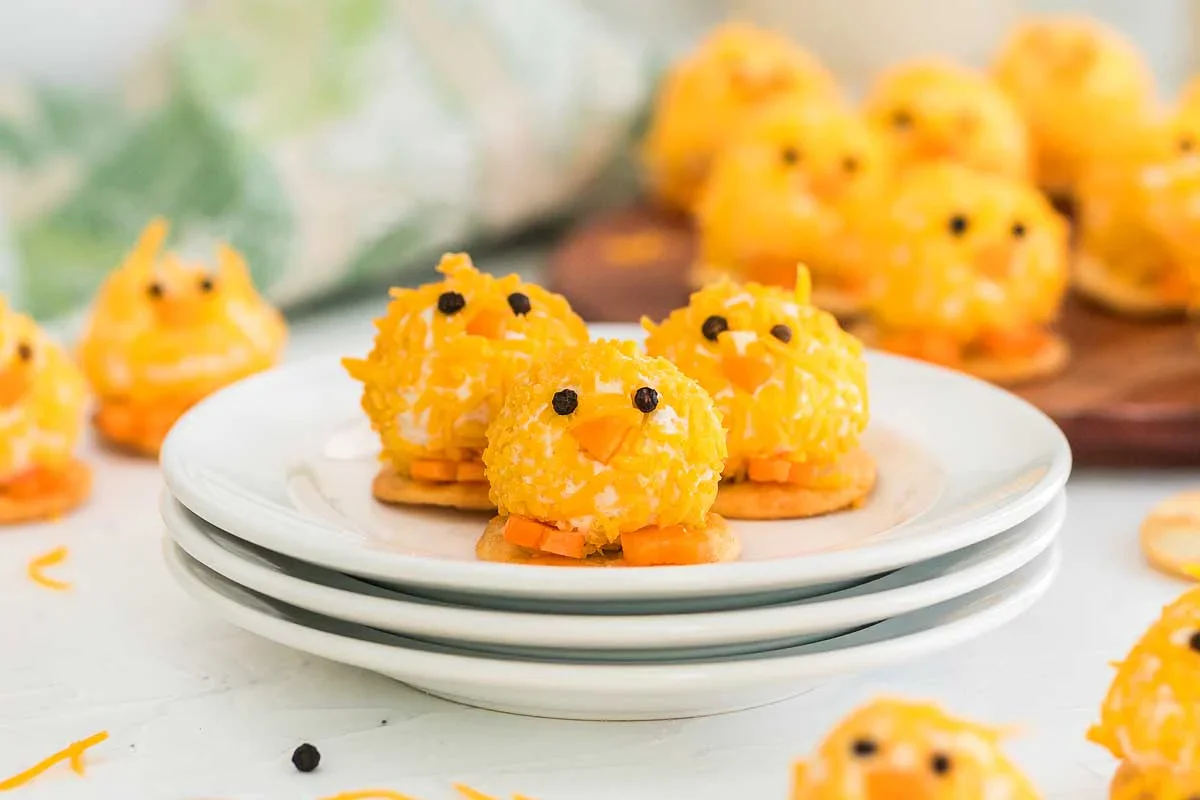 Three cheese balls decorated to look like chicks sit on stacked white plates, surrounded by more chick-shaped cheese balls on a light surface.