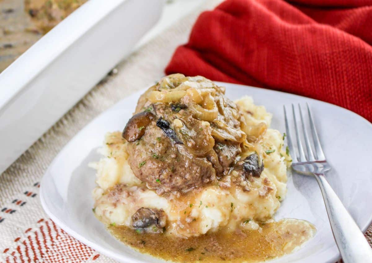 A serving of Salisbury steak with onions and brown gravy on mashed potatoes, on a white plate with a fork, next to a red cloth.