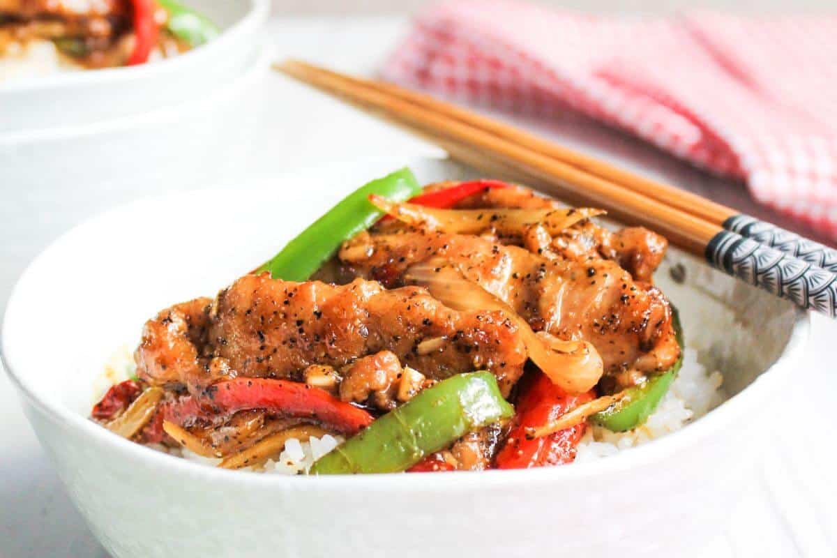 A bowl of white rice topped with stir-fried chicken, green and red bell peppers, onions, and sauce, with chopsticks resting on the bowl edge.