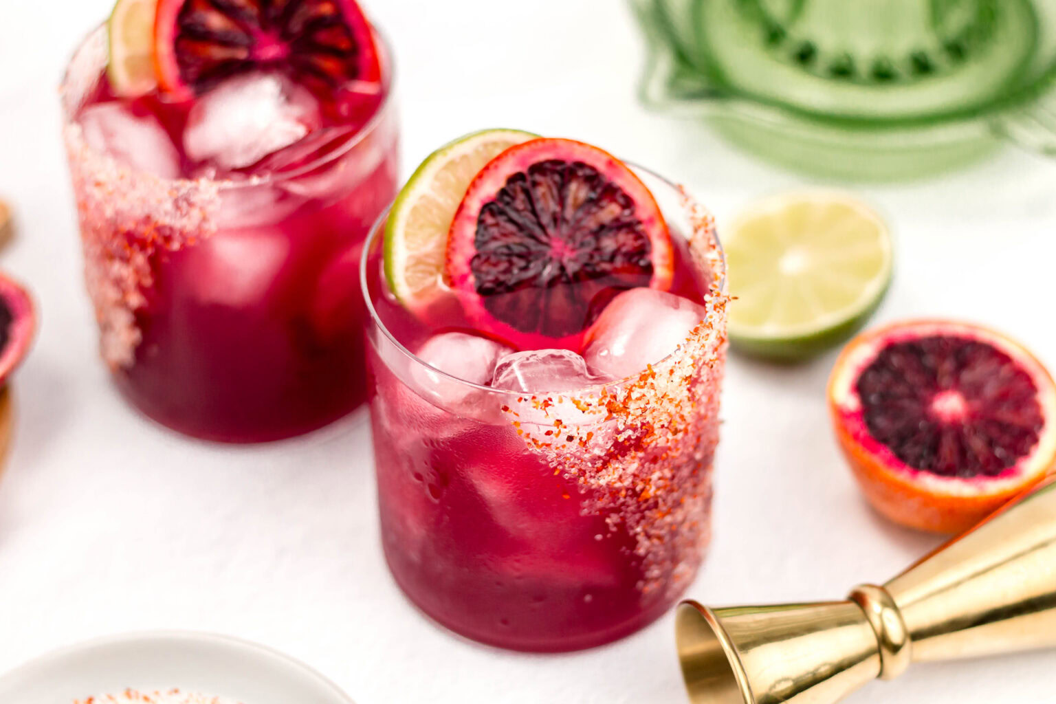 Two glasses of red cocktail with ice, lime, and blood orange slices, rimmed with chili powder, sit on a white surface next to fresh citrus fruit and a metal jigger.